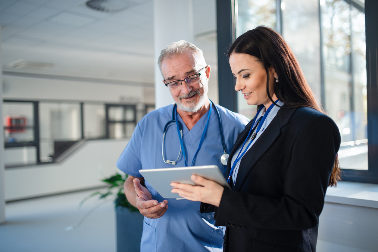 a woman wearing business attire and a doctor wearing scrubs looking at a tablet together