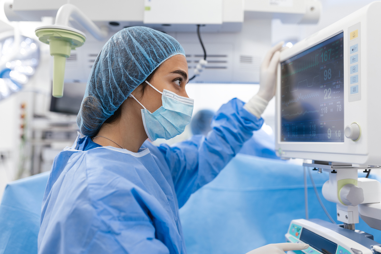 a doctor wearing scrubs, a mask, and a hairnet looking at a medical monitoring screen in a hospital