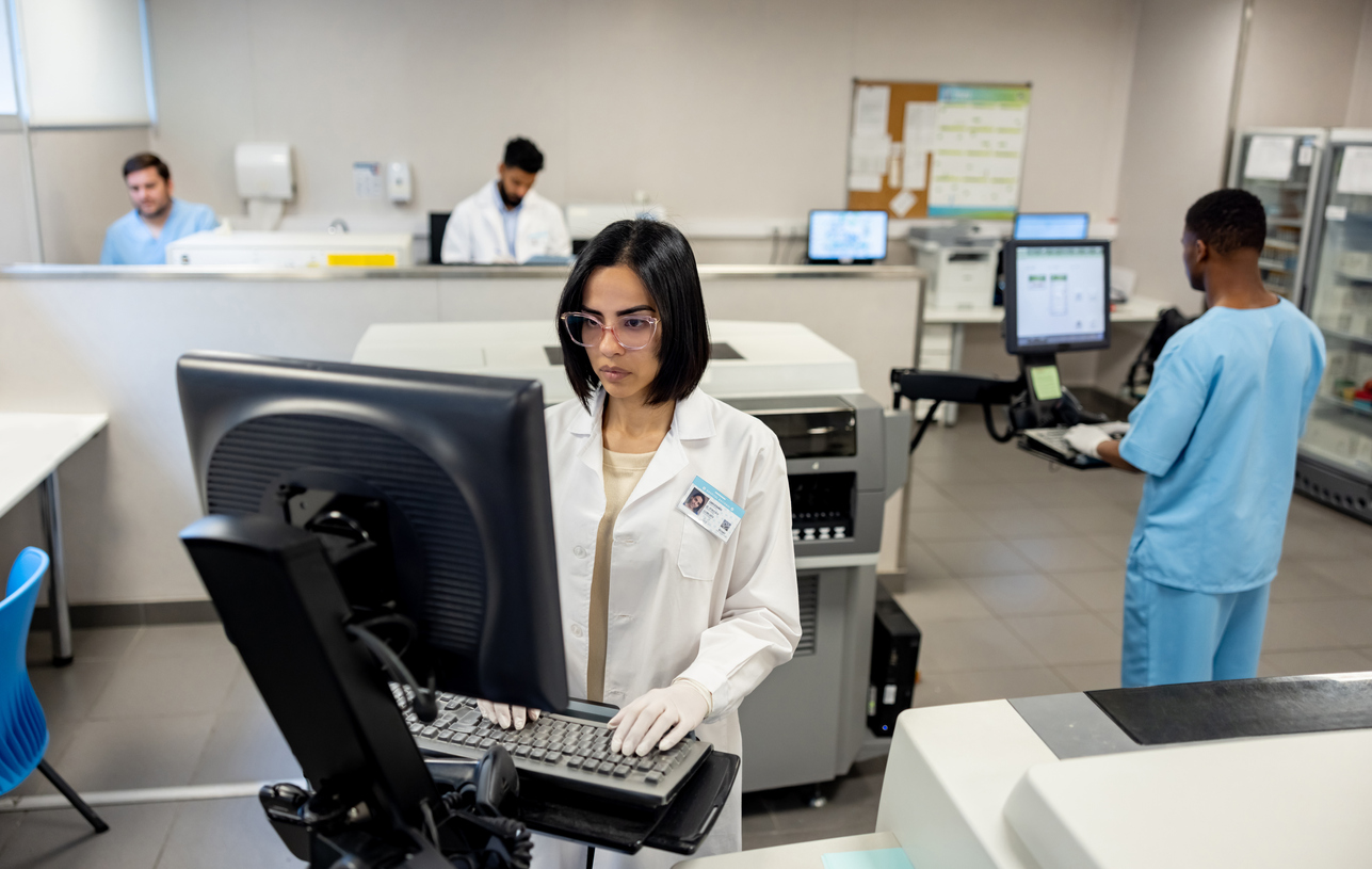 doctor in a lab coat typing on a computer in a hospital office surrounded by other medical professionals