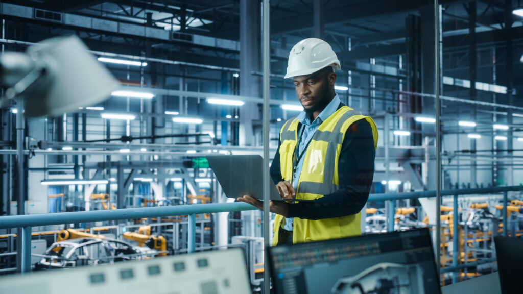 A manufacturing security engineer in a high-visibility vest and hard hat using a laptop to monitor and enforce network segmentation policies, ensuring IoT and OT device protection with Cylera’s cybersecurity platform in an industrial production facility.